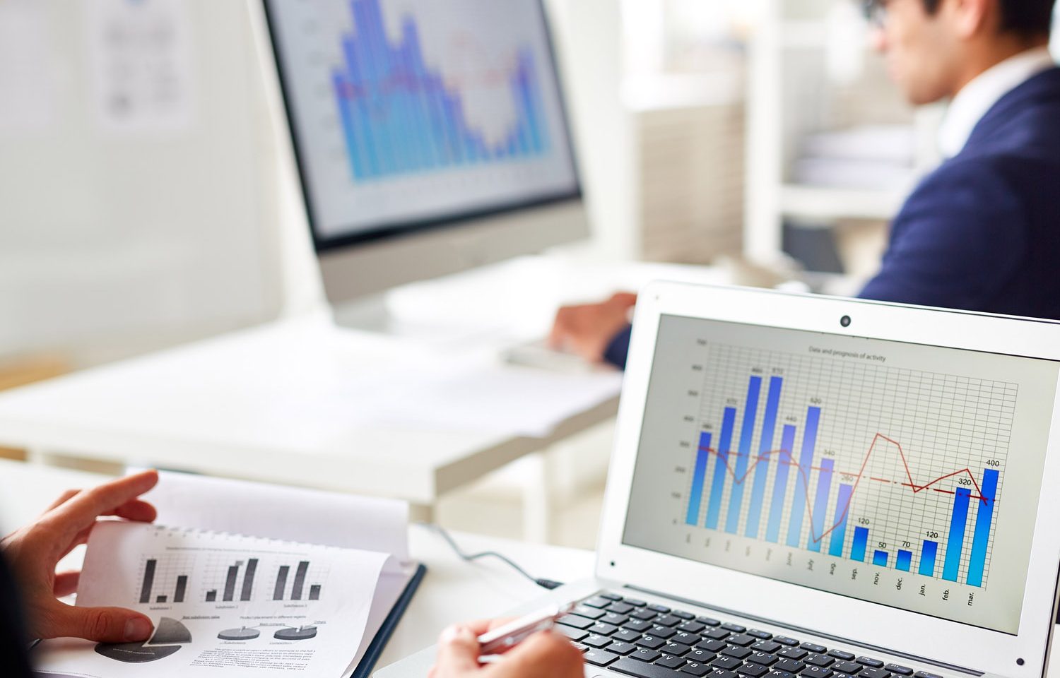 Person on laptop with graphs and paper with graphs, with person in background on computer with graphs and analytics